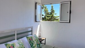 Ein Schlafzimmer mit einem Bett, Kissen, einem Nachttisch mit Lampe, einem Fenster mit Blick auf Bäume und einer Wandleuchte.