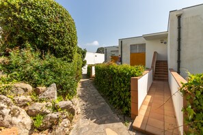 A narrow stone pathway flanked by greenery and white buildings with a staircase leading to an upper level.