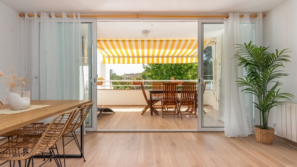 Un interior moderno con una mesa de comedor de madera, sillas de mimbre, una planta en maceta y un balcón con toldo a rayas amarillas.