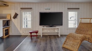 Sala de estar com televisão sobre suporte branco, uma poltrona de vime, um banco de madeira e uma área de cozinha com micro-ondas.