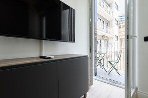Ein modernes Wohnzimmer mit einem Flachbildfernseher an einer dunklen Kommode, einer kleinen Fernbedienung auf der Kommode und Blick auf einen Balkon mit Metallgeländer und einem Stuhl.