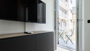 A modern living room with a flat-screen TV mounted on a dark cabinet, a small white remote, and a glass door leading to an outdoor balcony with a metal railing and two green folding chairs. - Via Niccol Tommaseo 4 - Appartamento 4 (Naples)
