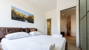 Cama bem arrumada com lençóis e almofadas brancas, um quadro de paisagem na parede e vista para outro quarto por uma porta aberta.