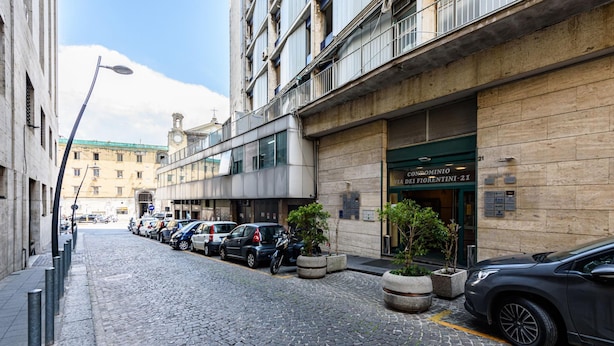 Uma rua de paralelepípedos com carros estacionados e um edifício com a placa 'CONDOMINIO VIA DEL FIORENTINO-21'.