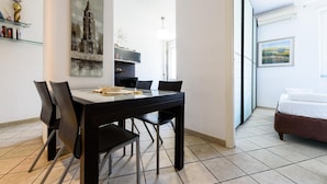 Uma zona de refeições moderna com uma mesa e cadeiras pretas, um quadro de um edifício na parede, e uma cama no quarto vizinho.