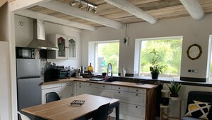A modern kitchen with a wooden table, two chairs, a sink, and a window with a view of greenery. - Le Majol , Gîte Rural au Coeur du Jura (La Chapelle-sur-Furieuse)