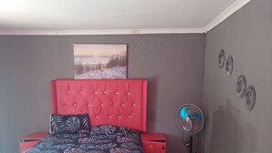 A bedroom with a red headboard, a bed with patterned bedding, a grey sofa, a small table, a standing fan, and a wall-mounted artwork. - Moholoholo lodge  (Bushbuckridge)
