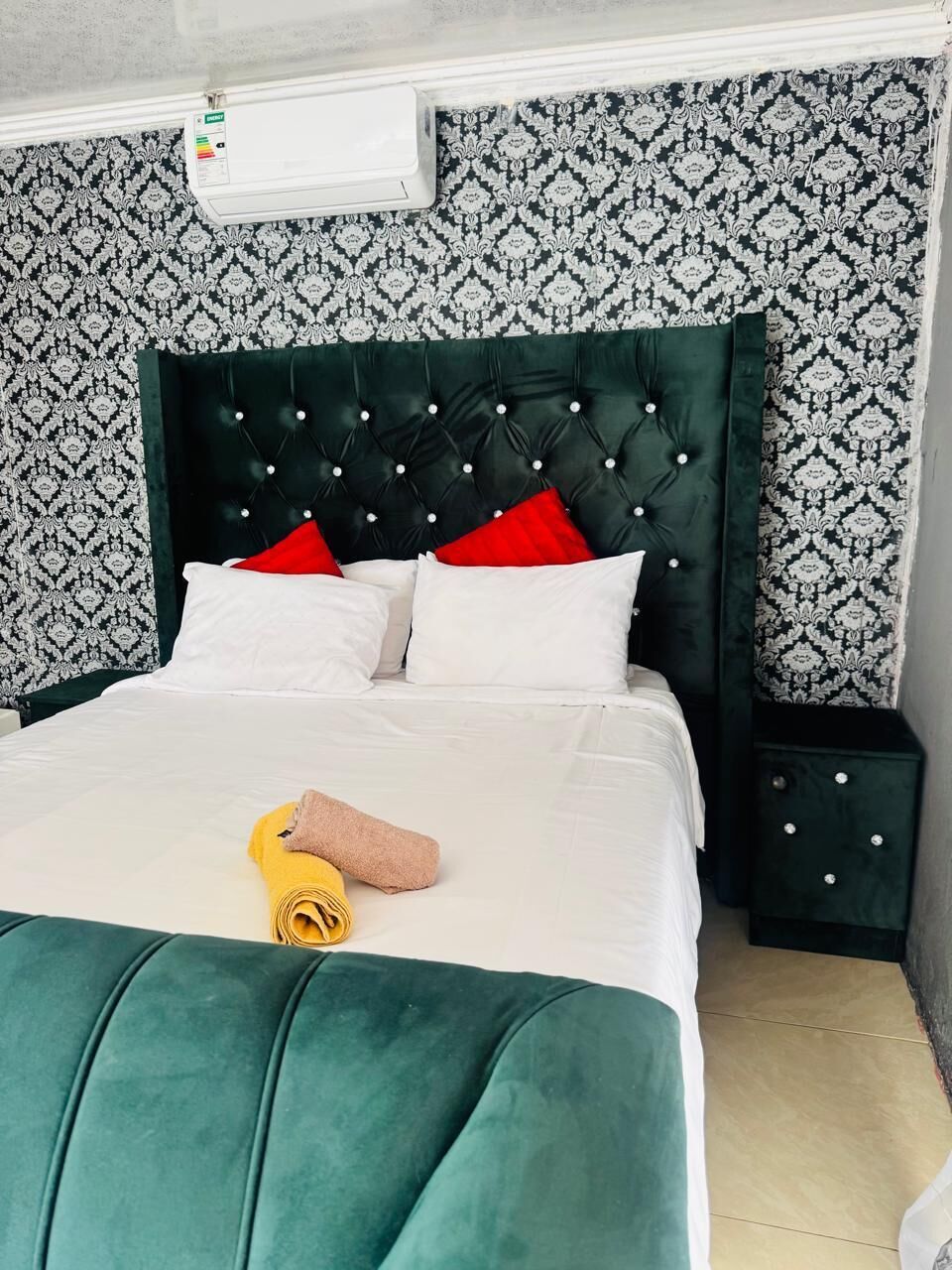 A bed with a tufted headboard, white bedding, and a rolled towel. There is a nightstand with a drawer and a small lamp. The room has patterned wallpaper and a tiled floor.