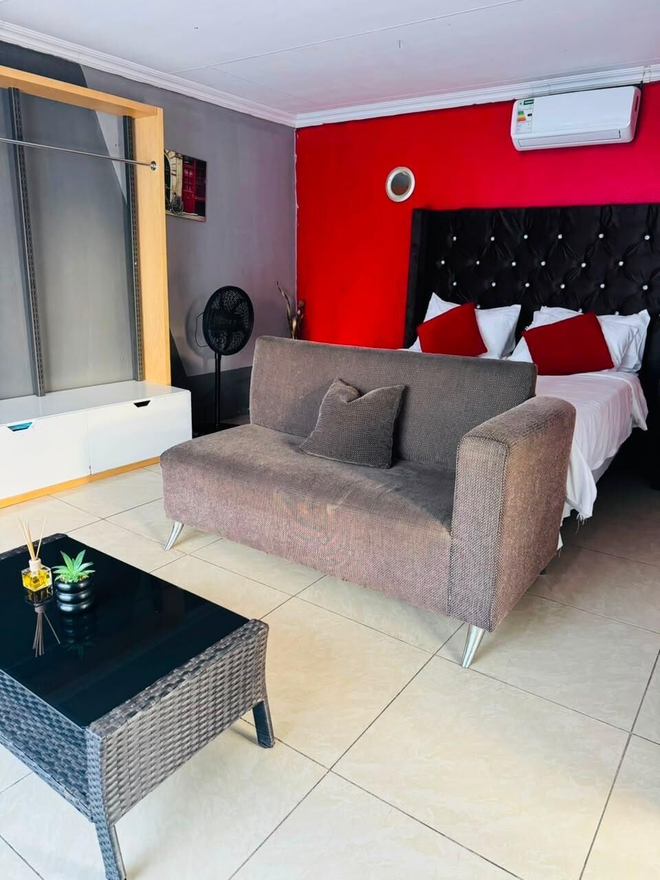 A room with a grey sofa, a black coffee table, a bed with red and white bedding, and a white air conditioning unit on the wall.