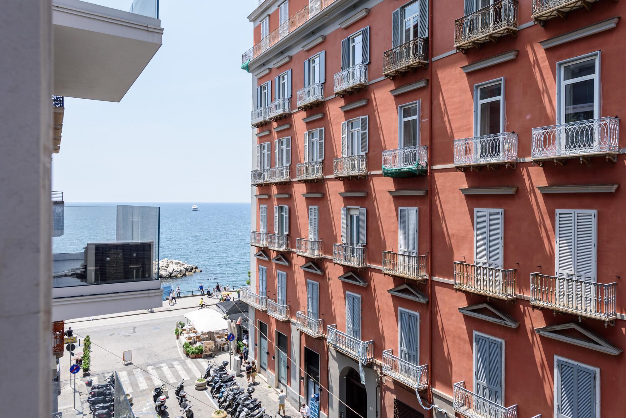 A multi-story building with balconies, a view of the sea, and a street with parked scooters.