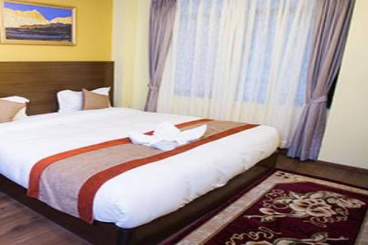 A hotel room with a wooden headboard, a bed with white linens and a red accent, a wooden desk, and a patterned rug.