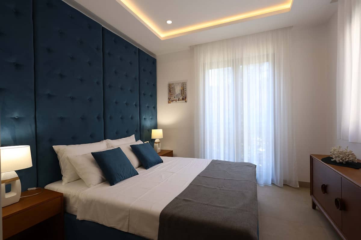 Une chambre d’hôtel moderne avec un grand lit, une tête de lit bleue, une table de chevet avec une lampe, une commode en bois et une grande fenêtre ornée de rideaux légers.