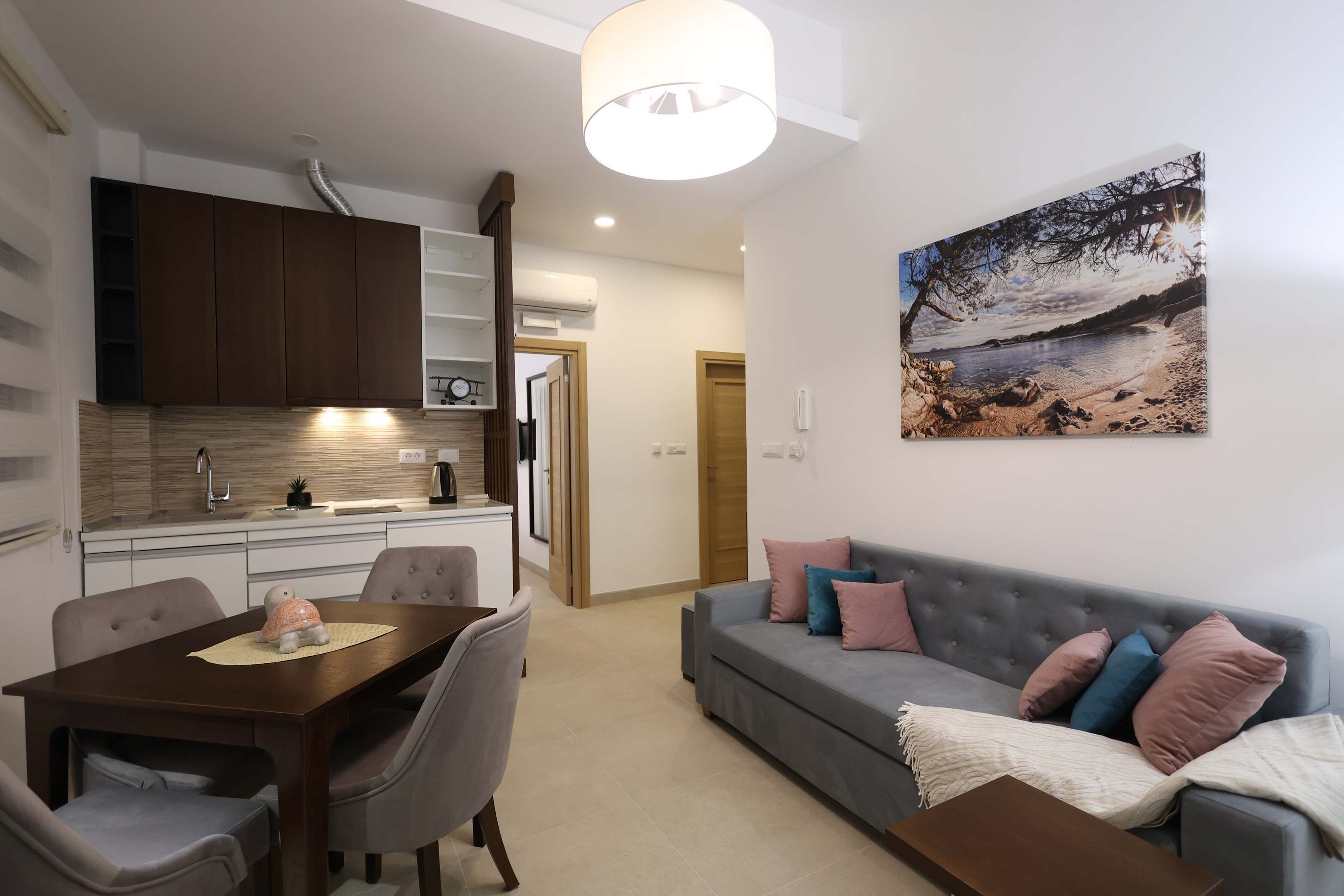 Una sala de estar moderna con un sofá gris, una mesa de comedor con sillas y una zona de cocina con armarios y un lavamanos.