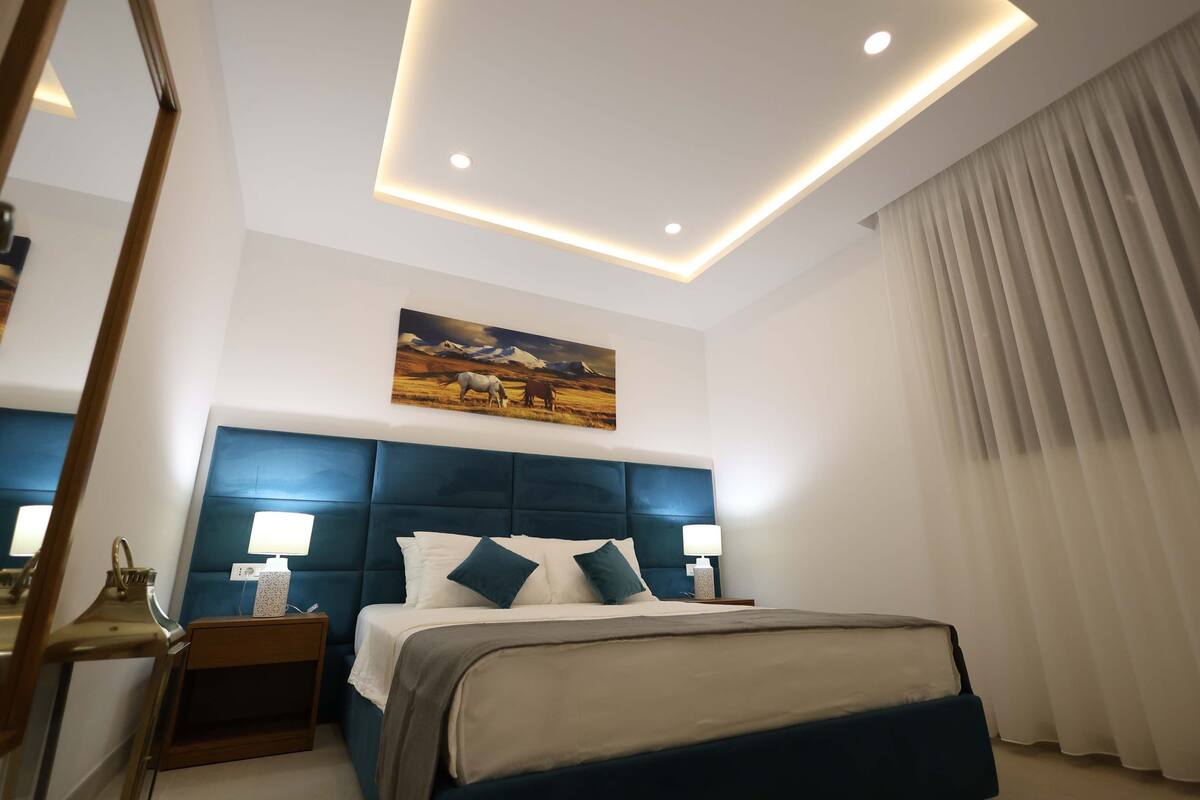 Une chambre d’hôtel moderne avec un grand lit, une tête de lit dotée d’un éclairage intégré, une table de chevet avec une lampe et un tableau au mur.