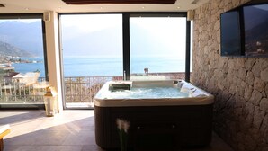 A modern room with a hot tub, a large window with a view of the sea, and a stone wall. - Bellezza Resort & SPA (Morinj)