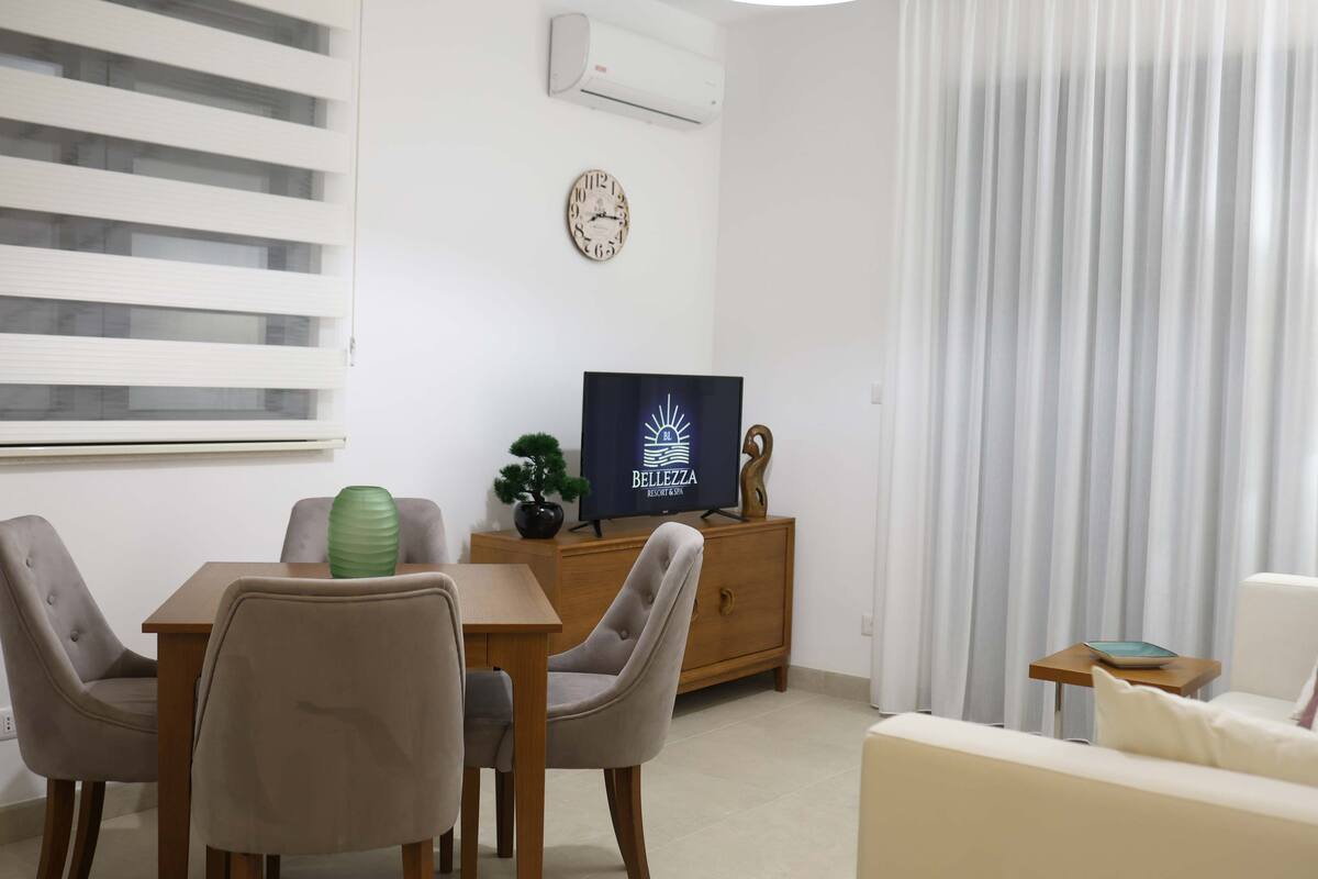 Un salon moderne avec un coin repas, comprenant un téléviseur, une horloge et une fenêtre avec des stores.