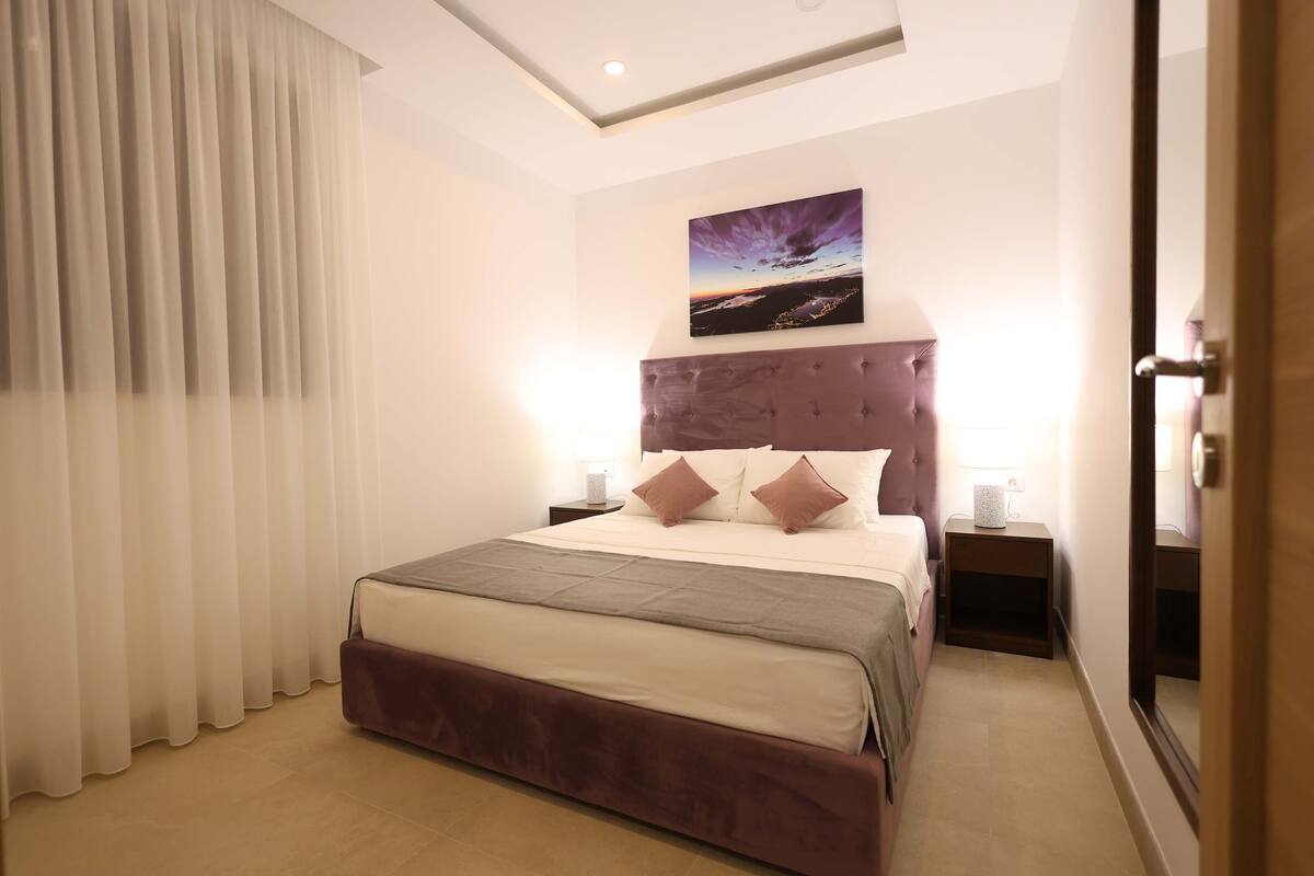 Une chambre d’hôtel moderne avec un grand lit, une tête de lit à motifs capitonnés, deux tables de chevet avec des lampes et une grande fenêtre avec des rideaux.