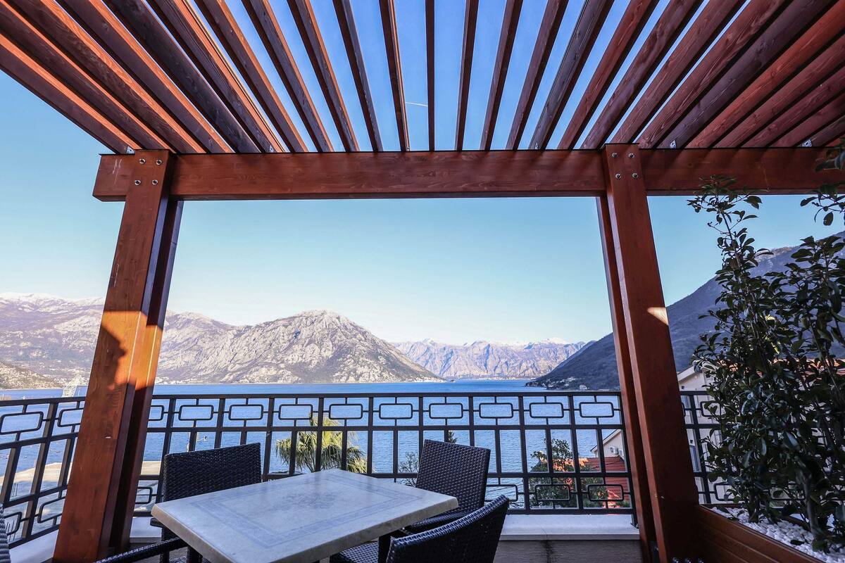 Une pergola en bois avec une table et des chaises, offrant une vue sur un paysage montagneux et une étendue d’eau.