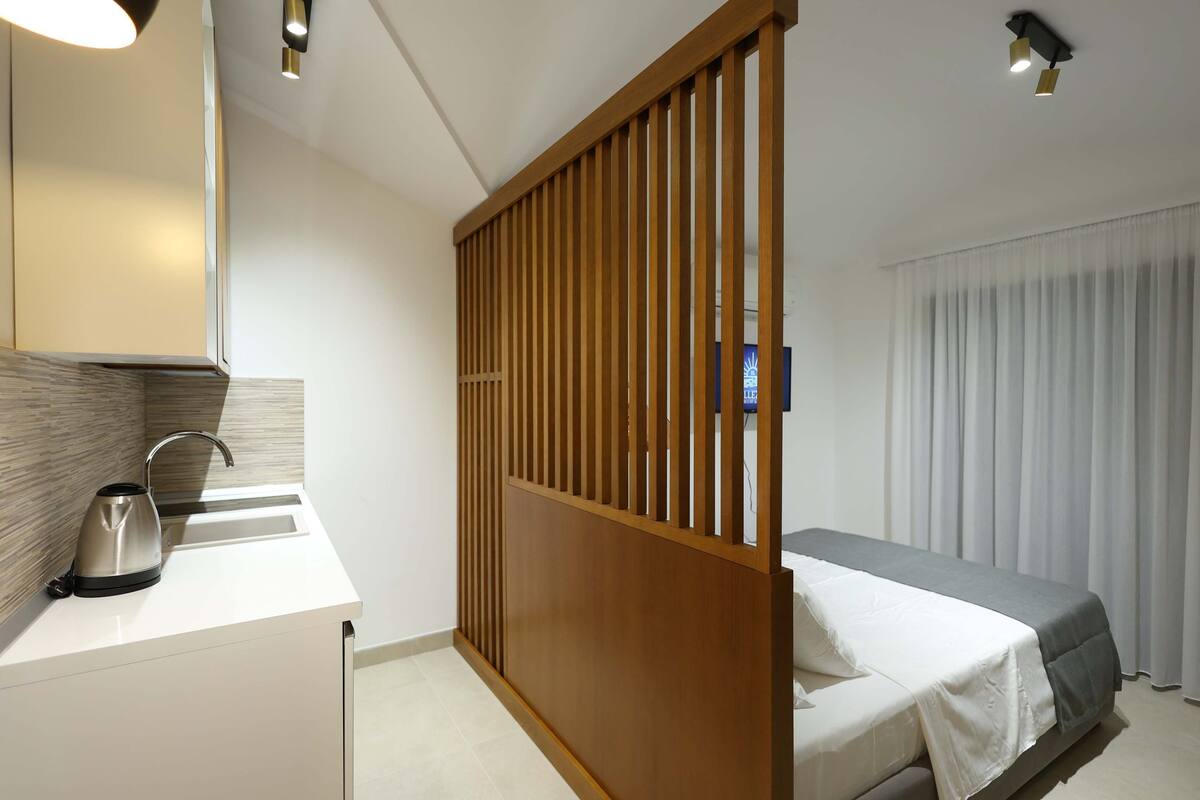 Une chambre d’hôtel moderne avec une cloison en bois, un lit recouvert d’une couverture grise, un lavabo blanc avec une bouilloire, et un téléviseur à écran plat fixé au mur.