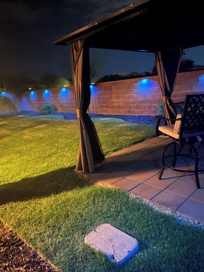 Uma área de pátio com um espaço de estar coberto, uma parede de tijolos e um jardim bem iluminado.