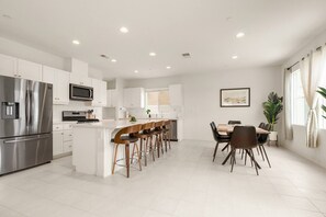 Una cocina moderna con gabinetes blancos, electrodomésticos de acero inoxidable, una isla central con taburetes y un comedor con mesa y sillas.