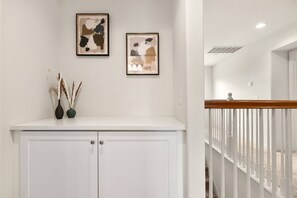 Un pasillo con una puerta blanca, dos cuadros abstractos enmarcados y un florero con plantas secas sobre una estantería.