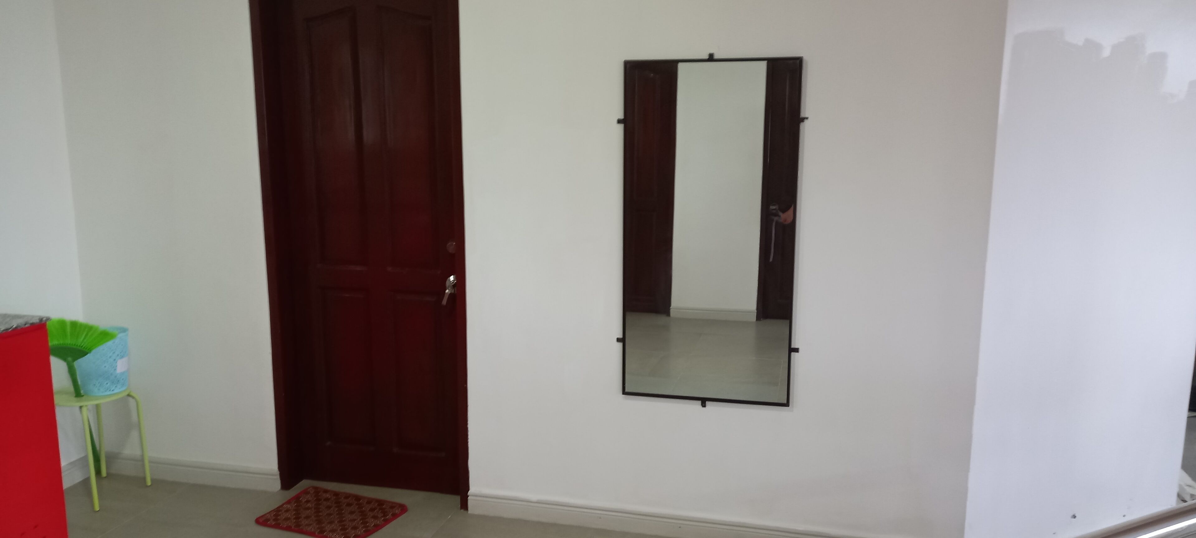 Een kamer met een houten deur, een spiegel aan de muur, een groene stoel en een rood voorwerp in de hoek.