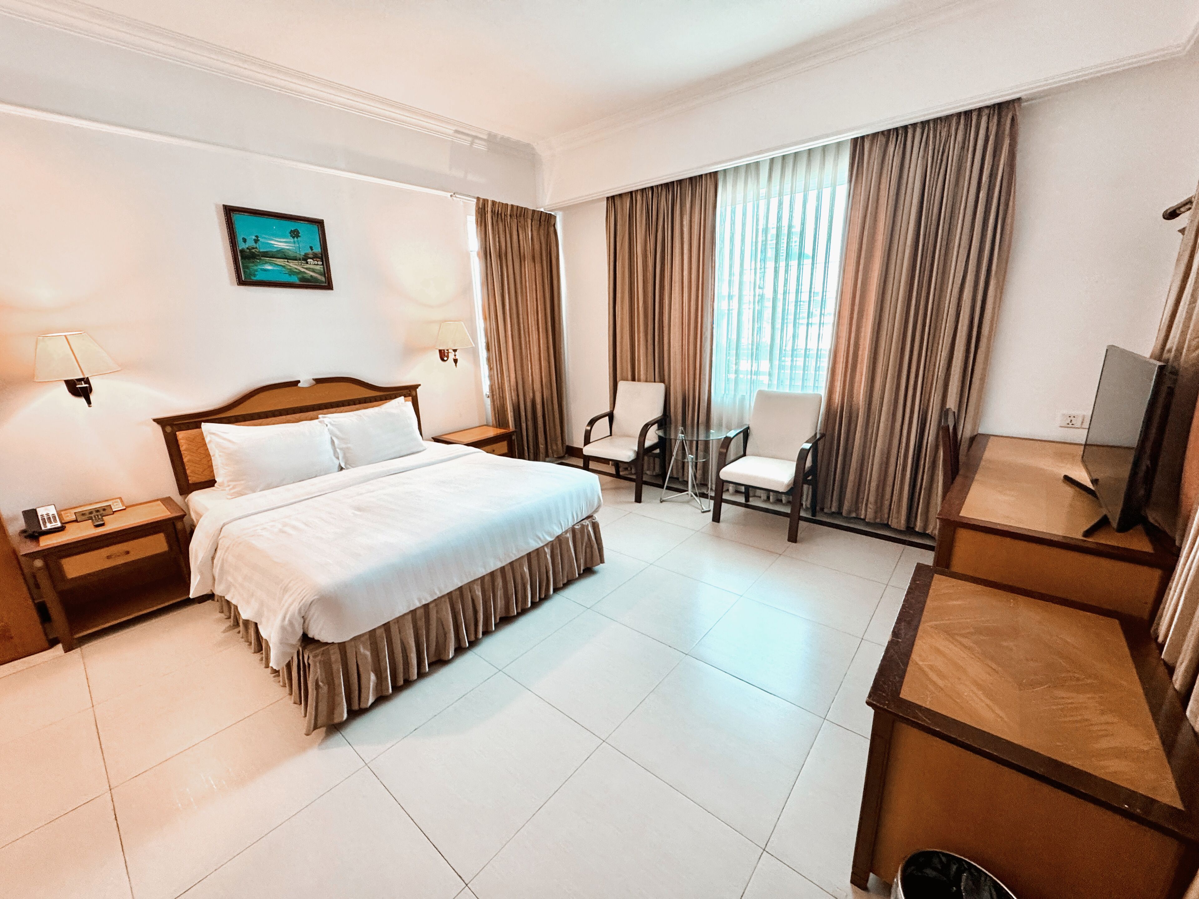 Quarto de hotel com cama, duas cadeiras, escrivaninha, televisão e um quadro na parede.