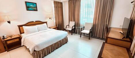 Quarto de hotel com cama, duas cadeiras, escrivaninha, televisão e um quadro na parede.