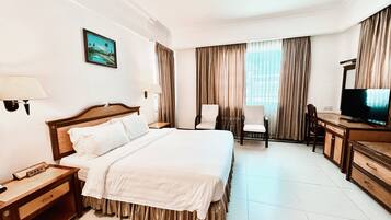 A hotel room with a large bed, a desk with a television, two chairs, and a window with curtains.