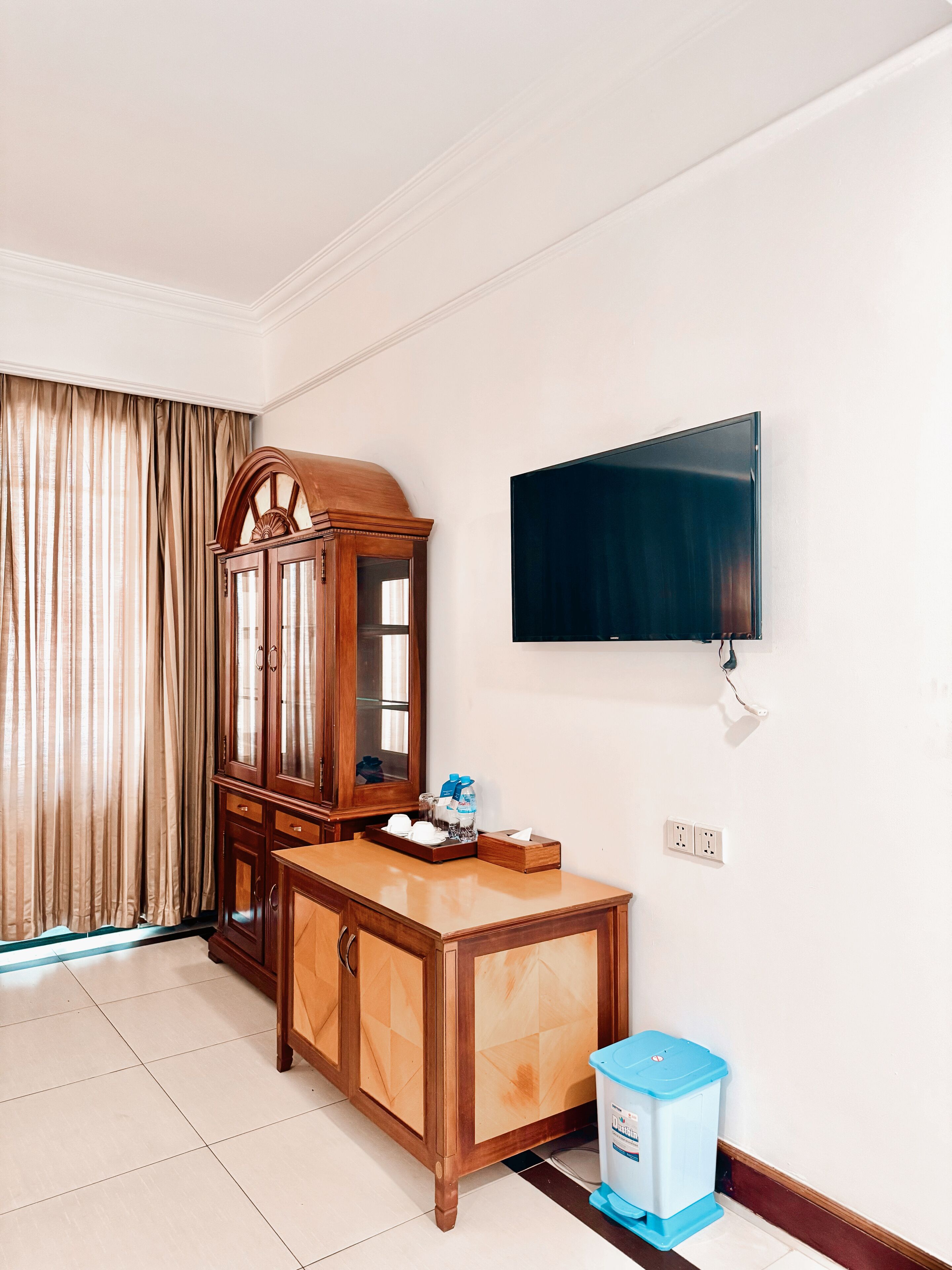 A hotel room with a wooden cabinet, a television mounted on the wall, a trash bin, and a window with curtains.