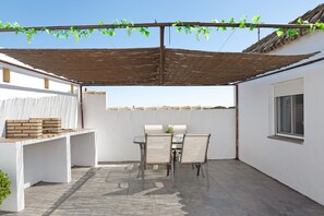 Une terrasse avec une table et des chaises, une pergola couverte de plantes grimpantes et un mur blanc avec une fenêtre.