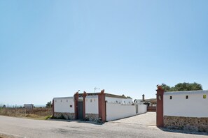 A gated entrance with a white wall and stone base, a clear sky, and a distant view of a building with a tiled roof. - Vacation Home "Casa Luype 2" with 2 Private Terraces, Shared Pool & Wi-Fi (Conil)