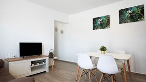 A modern living room with a flat-screen TV, a wooden entertainment unit, a dining table with white chairs, and two wall-mounted paintings.