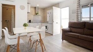 A modern kitchen with white appliances, a dining area with a white table and chairs, and a brown sofa in a living area.