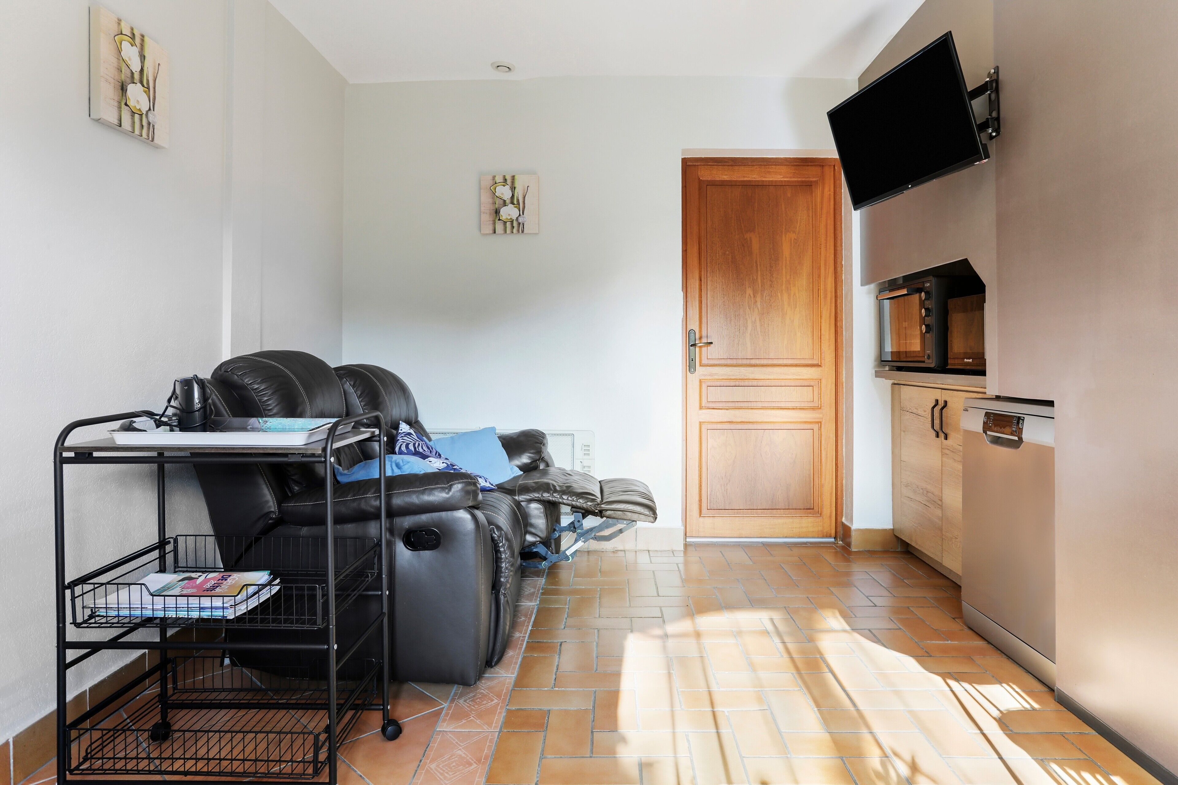 Un espace de vie compact comprenant un canapé, un petit meuble avec des livres, un four à micro-ondes et un téléviseur fixé au mur.