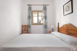 Un dormitorio con una cama de madera, una ventana con cortinas estampadas, una silla y una mesita de noche con una lámpara.
