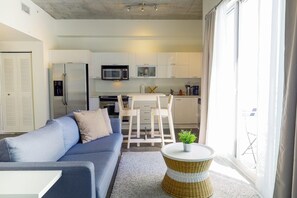 Cozy living space with a light blue sofa, wicker side table, and potted plant, adjacent to a kitchen area with white cabinetry and stainless steel appliances.
