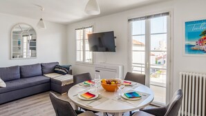 A modern living room with a grey sofa, a round dining table set with plates, glasses, and a fruit bowl. A flat-screen TV is mounted on the wall, and there is a large window with a view of buildings outside.