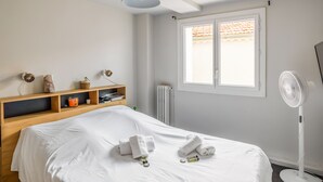 A neatly made bed with white linens and towels, a bedside table with a lamp, a shelf with books and a cup, and a window with a view of the outside.