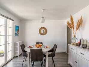 Uma sala de jantar com uma mesa redonda, quatro cadeiras, uma luminária de teto branca, um espelho decorativo e vista para o exterior através de grandes janelas.