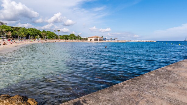 A beach with clear water, a concrete walkway, and buildings in the background.