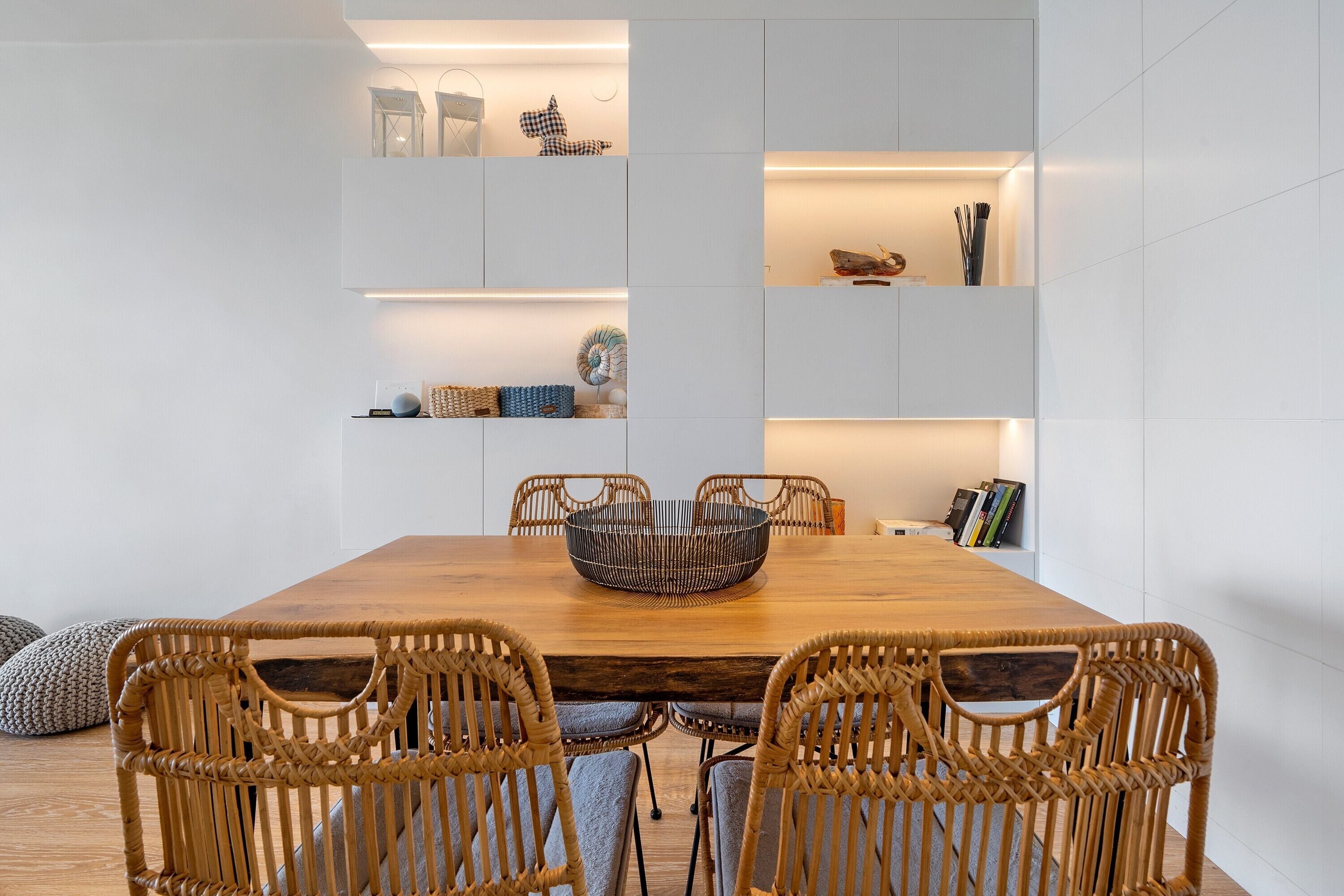 Un comedor con mesa de madera, sillas de mimbre y estanterías empotradas decoradas con objetos ornamentales.