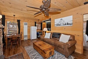 A cozy cabin interior with a wooden ceiling, a ceiling fan, a brown leather sofa, a dining table with chairs, and a large window. - Knotty By Nature By Destination Big Bear: Modern Cabin Decor! Spa! Pet Friendly! Close to Bear Mountain Ski Resort! (Big Bear Lake)