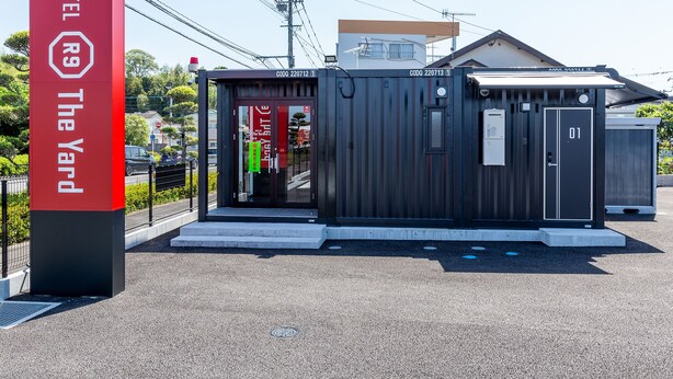 A compact hotel structure with a red sign indicating 'HOTEL The Yard'. It features a small entrance with a glass door, a visible room number, and a metal roof. - HOTEL R9 The Yard Izumi (Izumi)