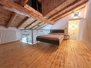 A loft bedroom with exposed wooden beams, a hanging bed, a decorative wrought iron railing, and a wall-mounted light fixture.