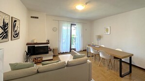 Ein modernes Wohnzimmer mit einer Couch, einem Fernseher, einem Esstisch mit Stühlen und einem großen Fenster mit Vorhängen.