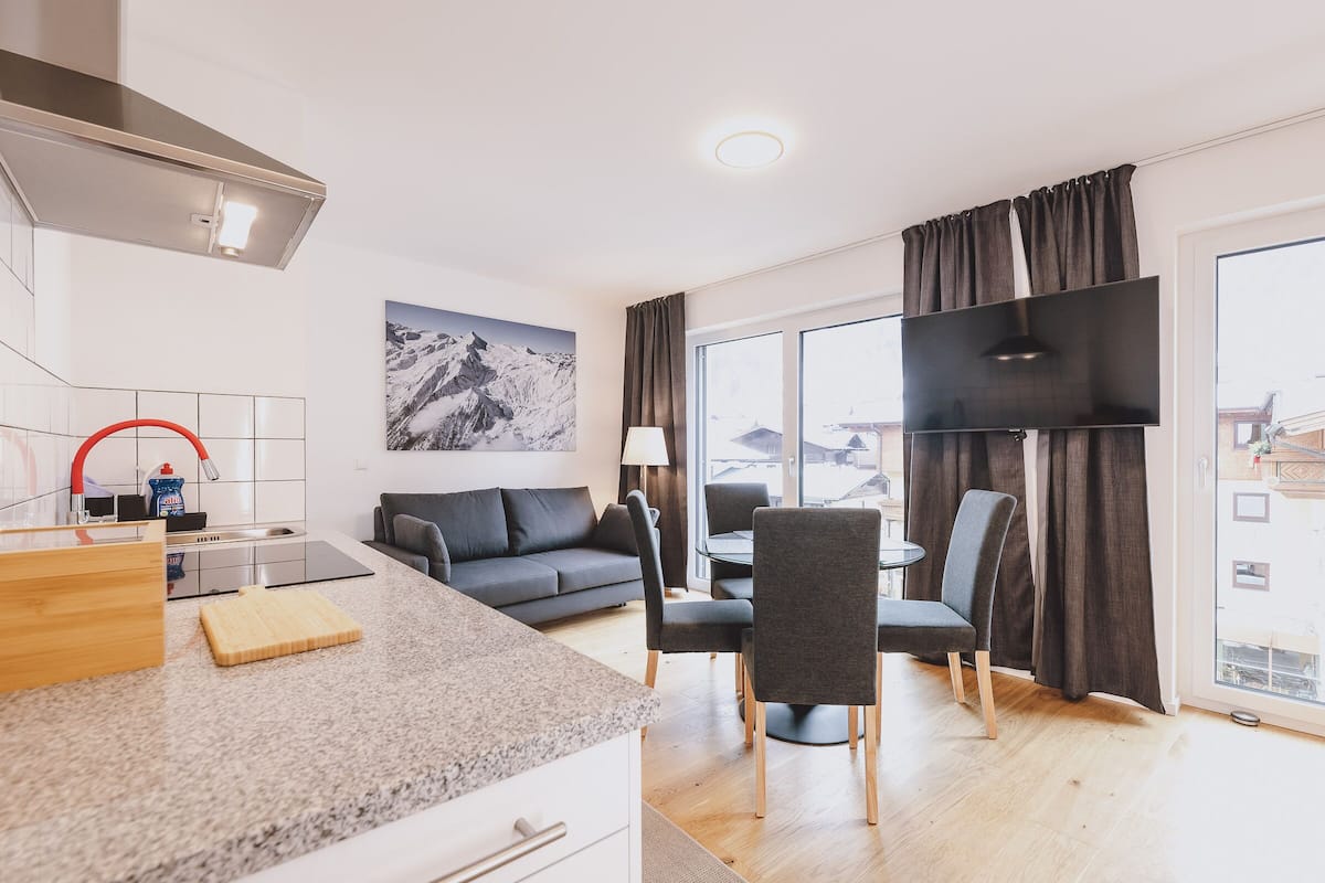 Moderni hotellihuone, jossa on keittotila, harmaa sohva, ruokapöytä tuoleineen ja suuri ikkuna, josta on näkymä rakennuksiin.