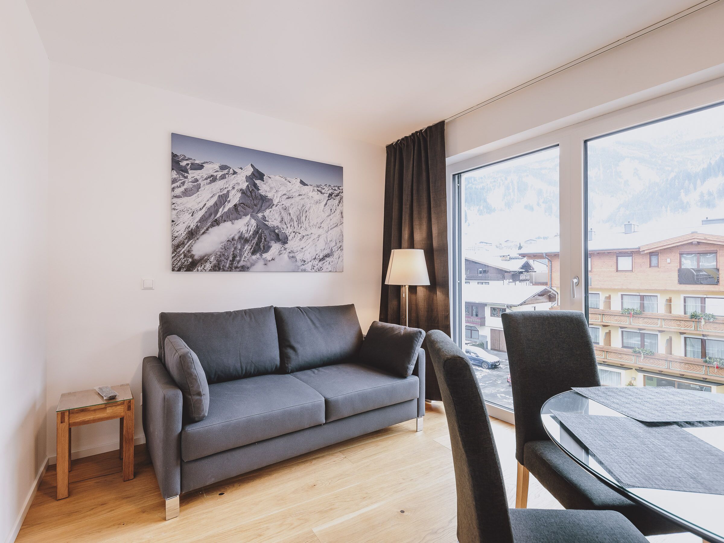 Een moderne woonkamer met een grijze bank, een glazen salontafel en uitzicht op een berglandschap door grote ramen.