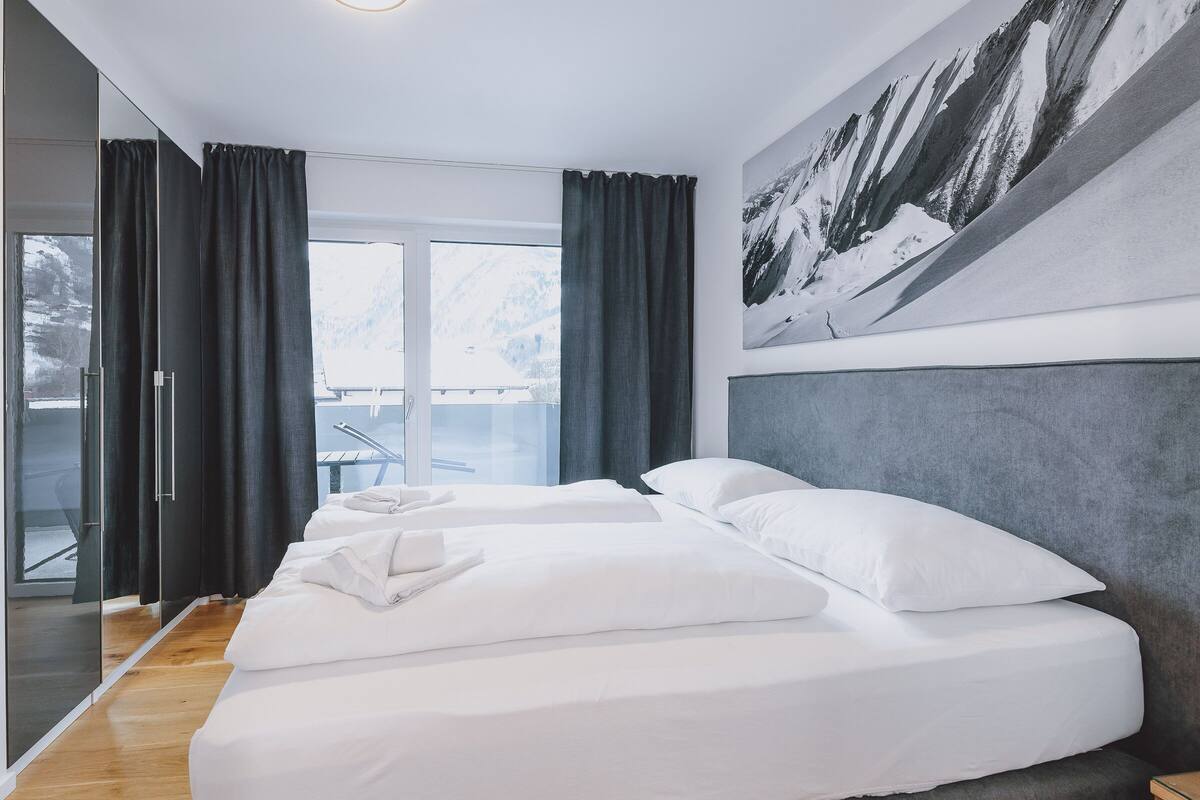 Hotellihuone, jossa on kaksi sänkyä, suuri ikkuna, jossa on verhot, vuoristomaisemaa esittävä taulu ja puulattia.